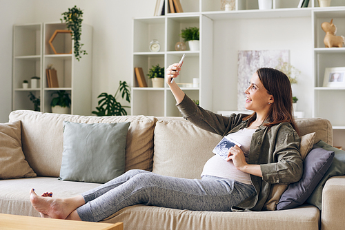 Happy pregnant woman with ultrasonic picture of her baby making selfie in front of smartphone camera while sitting on couch in living-room