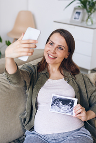 Cheerful attractive young pregnant woman sitting on sofa and photographing with ultrasound image of baby on smartphone