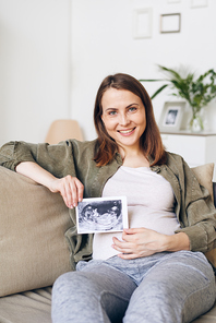 Portrait of happy attractive young woman in homewear sitting on sofa and holding fetal ultrasound image