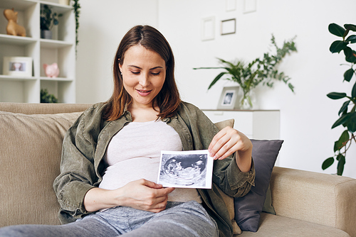 Contemporary pregnant woman showing you ultrasonic picture of future baby and looking at it while relaxing in home environment