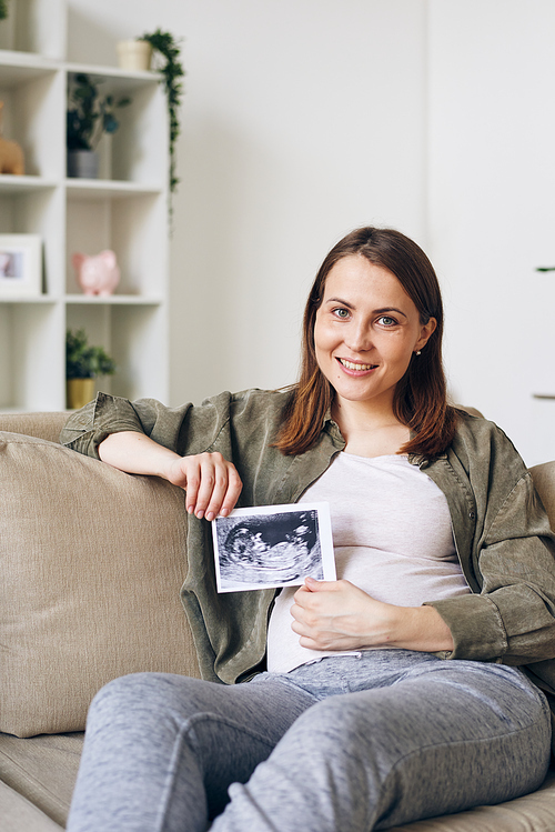Portrait of smiling beautiful young pregnant woman sitting in relaxed pose on sofa and posing with ultrasound photo of baby