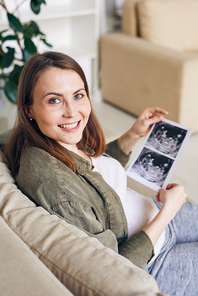 Portrait of positive young pregnant woman watching ultrasound photo of baby while sitting on sofa at home
