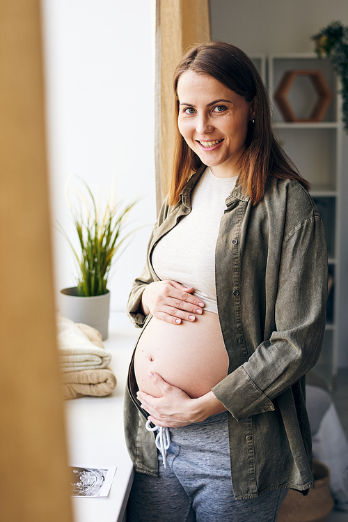 Portrait of happy young pregnant woman in shirt standing at window sill and hugging her belly