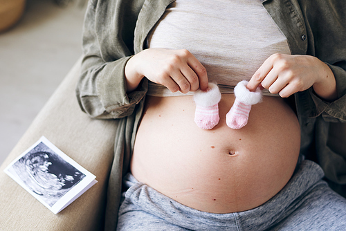 Close-up of unrecognizable pregnant woman holding cute socks for coming baby on her belly, fetal ultrasound image on elbow of sofa