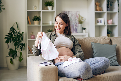 Happy young cross-legged pregnant woman looking at small shirt for future baby in her hands while sitting on couch in living-room