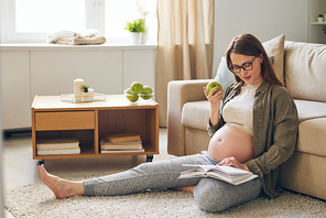 Young pregnant restful woman in casualwear reading book and eating green apple while sitting on the floor by couch at home