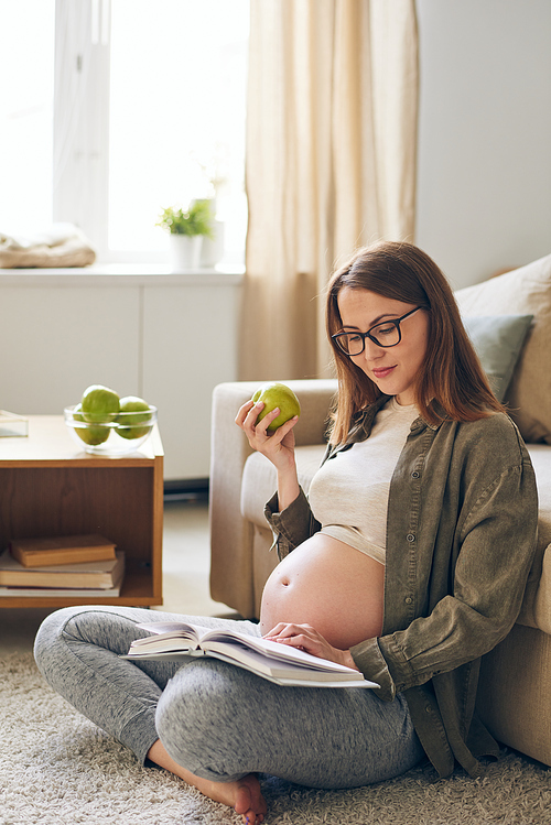 Content young pregnant woman in eyeglasses sitting with crossed legs on carpet and eating green apple while reading book at home