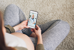 Above view of pregnant woman sitting with crossed legs on floor and using smartphone while talking to gynecologist via video chat