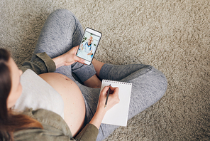 Top view of young pregnant female consulting with doctor in smartphone and making notes in notepad while sitting on the floor