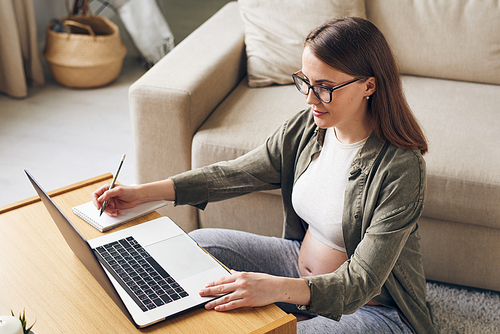 Content young pregnant woman in eyeglasses sitting at coffee table and using laptop while making notes about maternity at home