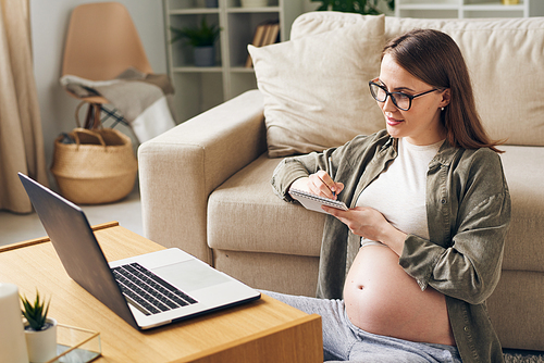 Happy pregnant woman making notes in notepad while sitting on the floor by couch in front of laptop during online medical consultation
