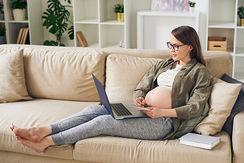 Attractive young pregnant woman in homewear sitting on sofa and reading article on laptop while making notes about maternity