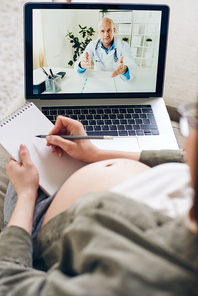 Contemporary pregnant female consulting with her doctor through video-chat and making notes while staying home on quarantine