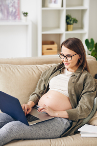 Content young pregnant woman in eyeglasses sitting on comfortable sofa and working with laptop using wi-fi at home
