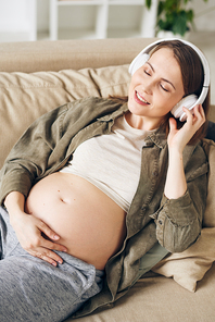 Young serene pregnant woman with headphones lying on couch, listening to calm music and relaxing in home environment