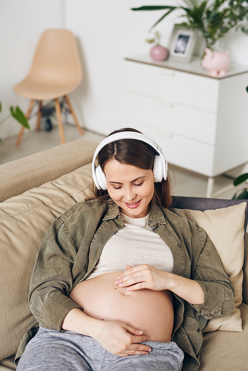 Happy young pregnant woman listening to relaxing music in headphones while lying on sofa in living room