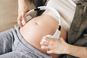 Hands of young pregnant female holding white headphones on both sides of her belly to let the baby listen to relaxing music