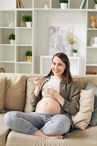 Portrait of positive young pregnant woman sitting with crossed legs on sofa and eating natural yogurt at home