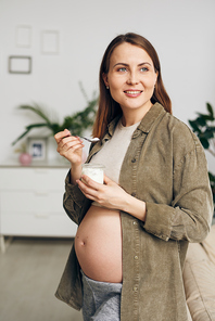Cheerful young pregnant female with toothy smile eating tasty yoghurt while standing by couch in living-room in front of camera