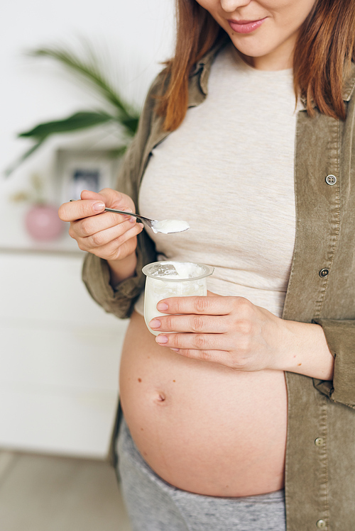 Close-up of content young pregnant woman with naked belly eating natural yogurt with spoon