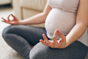Hand and leg bent in knee of young pregnant woman in activewear sitting in pose of lotus and meditating at home on the floor
