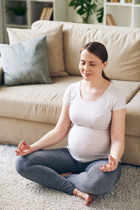 Young positive pregnant woman in activewear sitting in pose of lotus by couch on the floor and meditating while staying home