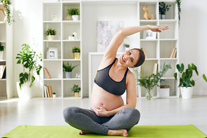 Portrait of smiling young pregnant woman in sports outfit sitting with crossed legs and bending aside with raised arm while practicing yoga at home