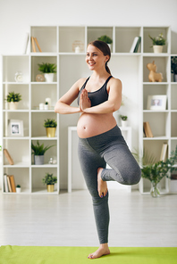 Happy young pregnant sportswoman standing on one leg on mat with her hands put together by chest during yoga training at home
