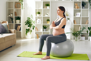 Young serene future mother in activewear sitting on fitball with her hands by chest during yoga training in home environment