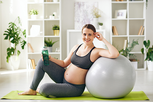 Young successful sporty pregnant woman with bottle of water leaning against fitball while sitting on mat in home environment