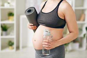Young active pregnant woman with rolled mat and bottle of pure still water standing in front of camera while going to do physical exercises