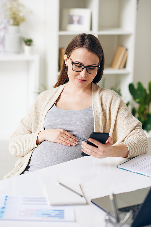 Young pregnant businesswoman in eyeglasses sitting at desk and checking phone messenger in office
