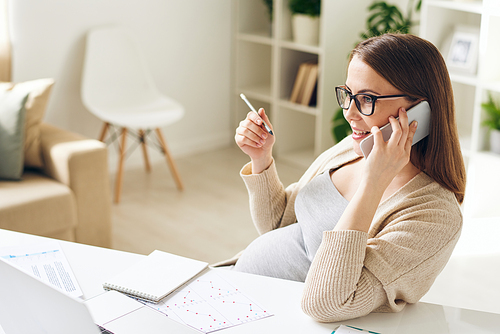 Confident young pregnant businesswoman in glasses sitting at desk with papers and answering client by phone in office