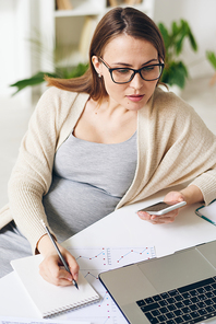Pretty pregnant analyst or broker using mobile gadgets while comparing online information with that in financial graph by workplace