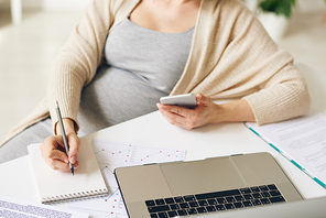 Close-up of unrecognizable pregnant businesswoman with smartphone in hand sitting at table and making notes about tasks while viewing data on laptop