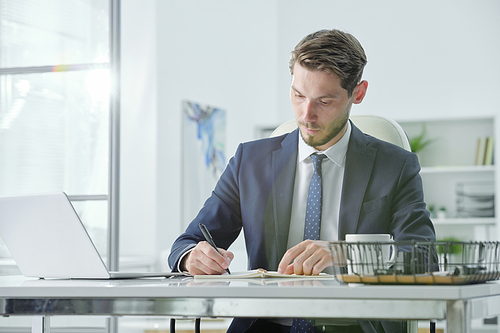 Concentrated handsome young businessman in gray suit sitting at desk and planning tasks while working in office