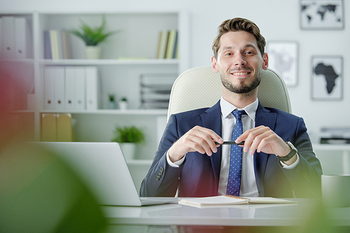Portrait of smiling confident young businessman with stubble sitting at office table with laptop and twisting pen