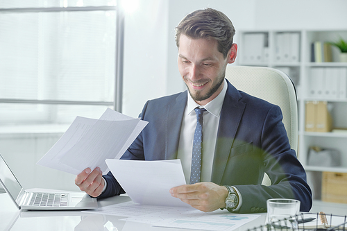 Satisfied young analyst sitting at desk and working with documents in office