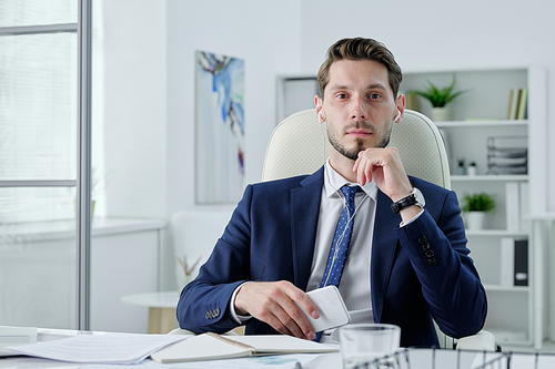 Portrait of serious business manager in earphones sitting at desk and holding modern phone in office
