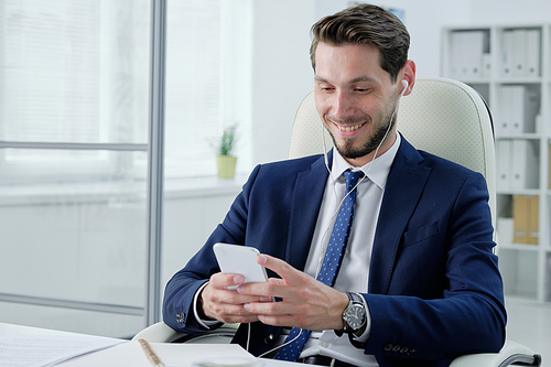 Positive young bearded businessman in earphones sitting on office chair and checking phone while listening to music