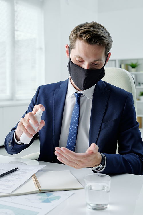 Modern young businessman in cloth mask sitting at table in office and reading contract while examining terms and conditions