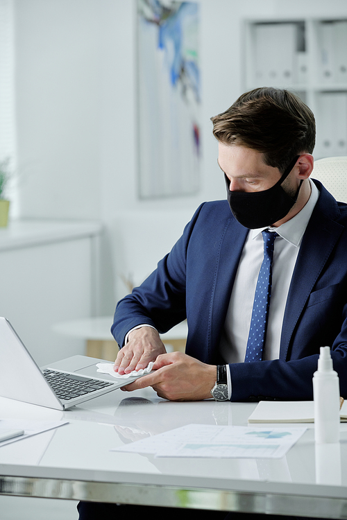 Young businessman in suit and facial mask sitting at desk and disinfecting laptop with wet napkin in office