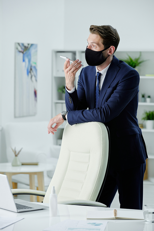 Young businessman in suit and facial mask sitting at desk and disinfecting laptop with wet napkin in office