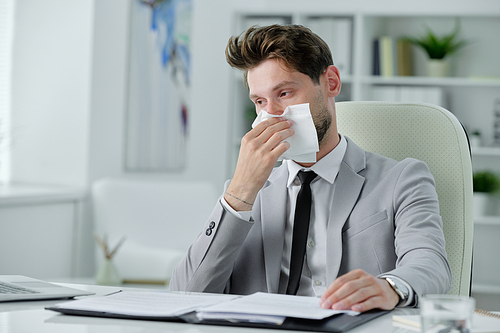 Ill young businessman sitting at desk and rubbing runny nose while working with papers in office, cold and flu concept