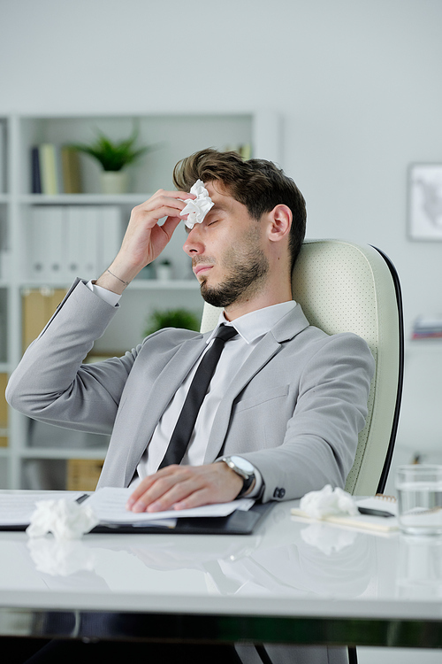 Ill young businessman sitting at desk and rubbing runny nose while working with papers in office, cold and flu concept