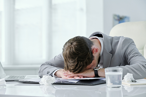 Exhausted young businessman lying on table with papers and crumpled napkins while suffering from headache