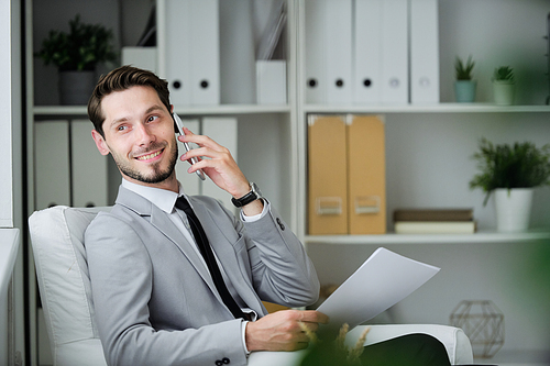 Cheerful successful young businessman with beard sitting in comfortable armchair in office and viewing contract while communicating by phone