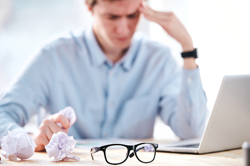 Close-up of young businessman crumpling papers while having not ideas for project in office, focus on eyeglasses in foreground