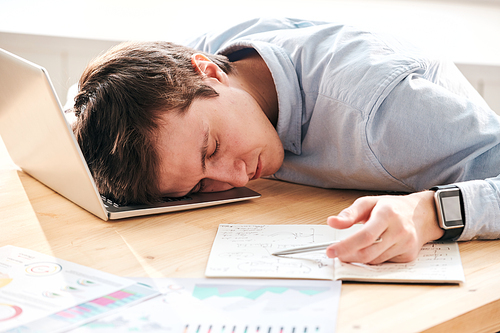 Young tired man exhausted with calculations lying on laptop while sleeping at workplace