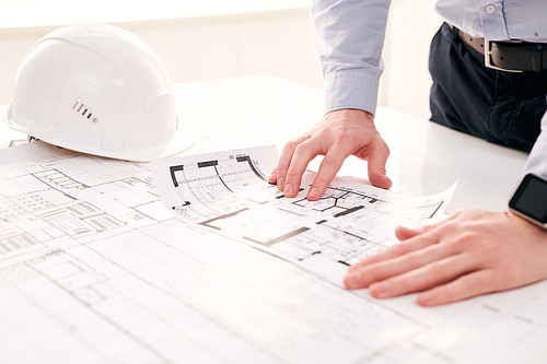 Close-up of unrecognizable engineer standing at table with hardhat and examining blueprint